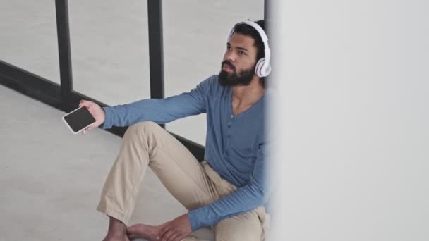 自宅のリビングルームでヘッドフォンをしながら 夢のような穏やかなアフリカ系アメリカ人の男が彼のスマートフォンを持っている — ストック動画