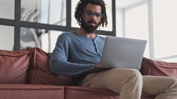 一位戴着眼镜的严肃的非洲裔美国年轻人正坐在自家客厅里用他的银色笔记本电脑 — 图库视频影像
