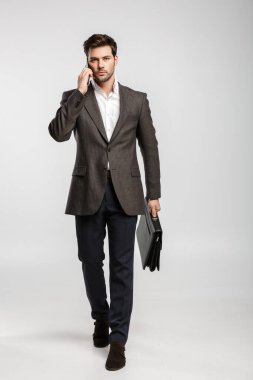 Ceketli genç başarılı iş adamının görüntüsü beyaz arka planda izole edilmiş bir çantayla yürürken cep telefonuyla konuşması