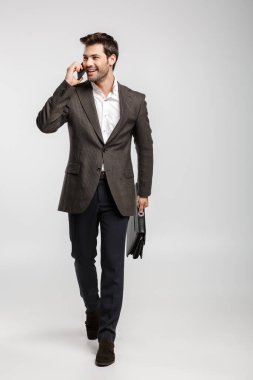 Ceketli gülümseyen bir iş adamının beyaz arka planda çantasıyla yürürken cep telefonuyla konuşması.