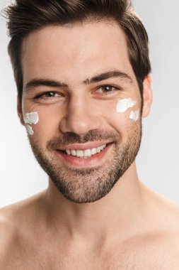Yüz kremi sürmüş, gülümseyen ve beyaz arka planda izole edilmiş kameraya bakan neşeli yarı çıplak adamın fotoğrafı.