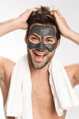 Yüzünde kozmetik kil maske ve beyaz arka planda izole edilmiş kameraya gülümseyen mutlu yarı çıplak adamın fotoğrafı.