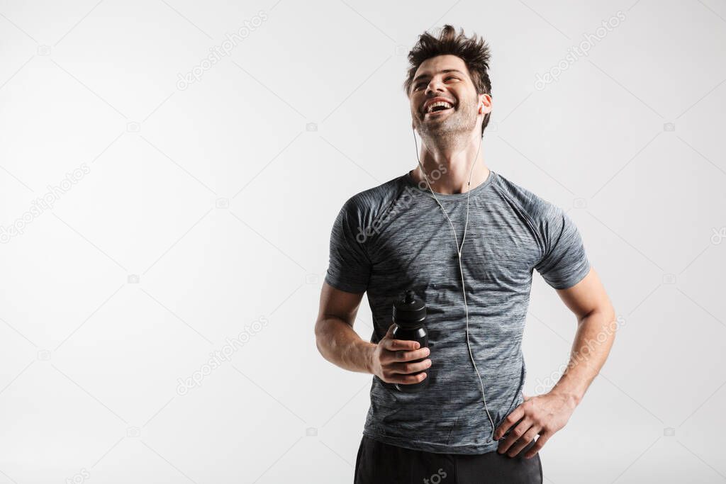 Image of young laughing man using earphones and holding water bottle while working out isolated over white background