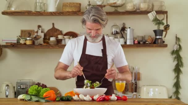Ein Glücklicher Älterer Mann Schürze Mixt Salat Einer Gemütlichen Küche — Stockvideo