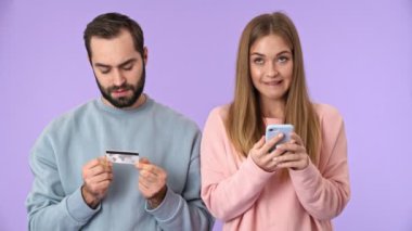 Neşeli sarışın kadın, mor arka planda kredi kartı tutan sakallı adamın yanında dururken akıllı telefonuyla alışveriş yapıyor.