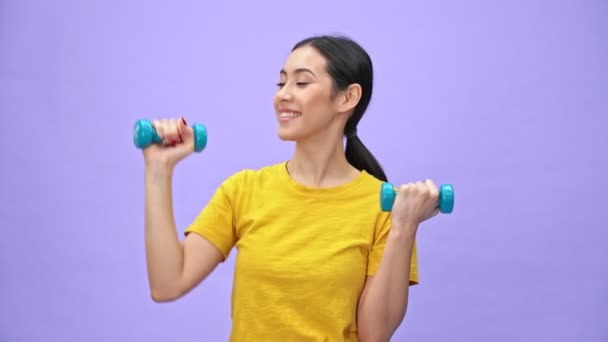 幸せなかわいいですスポーツ女性で黄色Tシャツ行います運動とともにダンベルその後 上のカメラを見ますバイオレット背景 — ストック動画