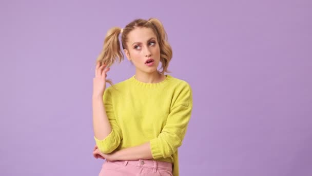 一个穿着黄色毛衣的迷人的金发姑娘厌倦了说话 她在一个紫色背景的孤立的工作室里扭动着手指上的头发 — 图库视频影像