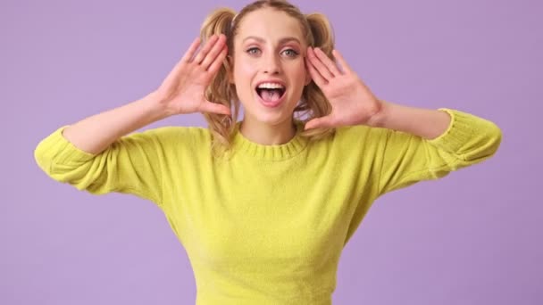 一个穿着黄色毛衣的金发美女对着摄像机笑着 并在一个紫色背景的孤零零的工作室里做鬼脸 — 图库视频影像