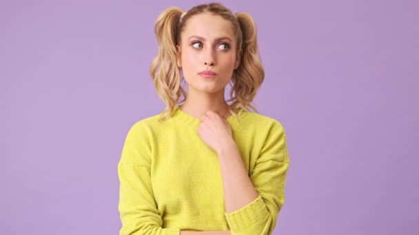 一个穿着黄色毛衣的漂亮金发姑娘把她的手放在毛衣旁边 仔细地听着这个建议 仔细地考虑了一下 然后同意了 在一个紫色背景的孤立的工作室里摇着头 — 图库视频影像