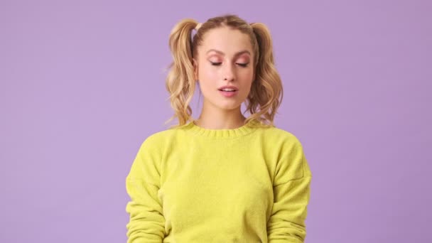 黄色のロマンチックなセーターの素晴らしい女の子は単にウィンクし 笑顔でスタジオに隔離された距離の誰かで紫色の背景 — ストック動画