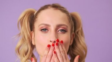 Kameraya bir öpücük yollayan hoş, genç, utangaç bir sarışının portresi. Mor bir arka planda, izole edilmiş bir stüdyoda parmaklarıyla saçlarını büküyor.