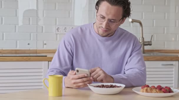 在家里吃早餐的时候 用智能手机对着眼镜笑英俊的男人 — 图库视频影像