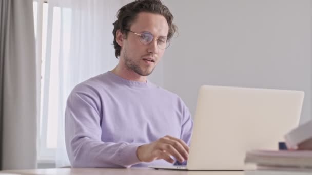 幸せなハンサムな男で眼鏡でビデオコールによってラップトップコンピュータ上で座っている間にテーブルに家 — ストック動画