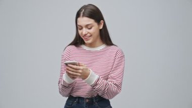 Neşeli, çekici esmer kadın akıllı telefon kullanıyor ve gri arka planda eğleniyor.