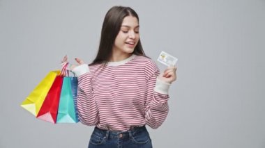 Elinde paketler ve kredi kartıyla gülümseyen çekici esmer kadın gri arka planda anın tadını çıkarıyor.