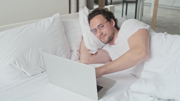 ベッドルームでベッドの上に横たわっている間 ラップトップコンピュータを使用して眼鏡でハンサムな男を笑顔 — ストック動画
