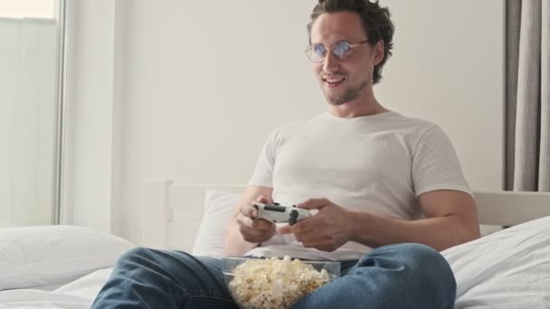 自宅でソファの上にポップコーンと座っている間 ゲームパッドでコンソールで遊んで眼鏡をかけてハンサムな男 — ストック動画
