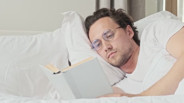 Sorrindo Homem Bonito Óculos Lendo Livro Enquanto Deitado Cama Quarto — Vídeo de Stock