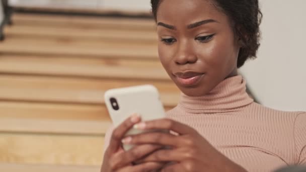 一位心平气和 可爱的年轻非洲女子正坐在家里的楼梯上 一边用她的智能手机 — 图库视频影像