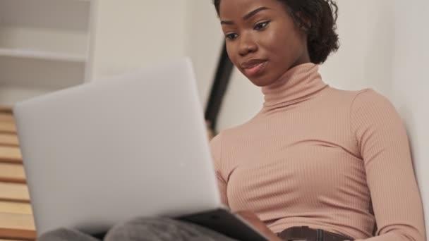 Серйозна Молода Африканка Дивиться Щось Своєму Ноутбуці Сидячи Сходах Вдома — стокове відео