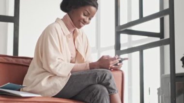 Memnuniyet duyan genç Afrikalı bir kadın cep telefonundan bir şey izliyor. Evde çalışırken onu yatay olarak tutuyor.