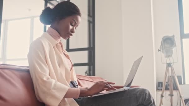 一位年轻貌美的非洲女性室内设计家坐在客厅的沙发上 一边用她的笔记本电脑 — 图库视频影像