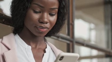 Güzel gülümseyen Afro-Amerikan bir iş kadını ofisinde akıllı telefonunu kullanıyor.