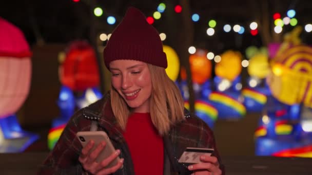 快乐的金发姑娘穿着秋天的衣服 用智能手机和信用卡在霓虹灯照明的背景下付账单 — 图库视频影像