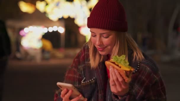 笑顔若いですブロンドの女の子身に着けています秋の服スマートフォンを使用して ネオンの照明の背景にサンドイッチを食べる — ストック動画