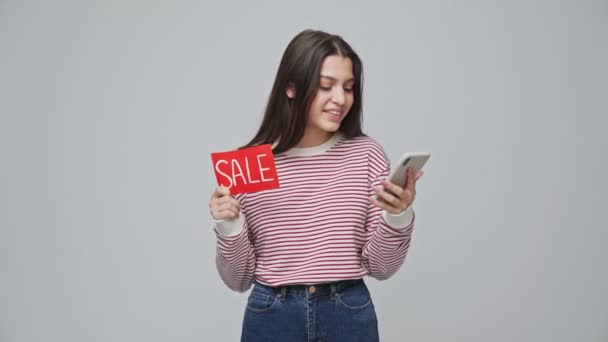 笑顔魅力的なブルネット女性保持ネームプレート販売ながら使用スマートフォン以上グレーの背景 — ストック動画
