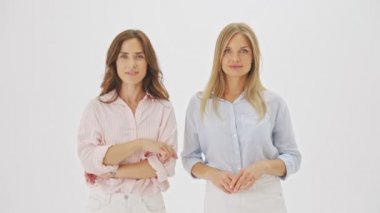 İki çekici kadın kameraya bakarak gizli bir jest yapıyor. Stüdyonun beyaz arka planında izole bir şekilde duruyorlar.