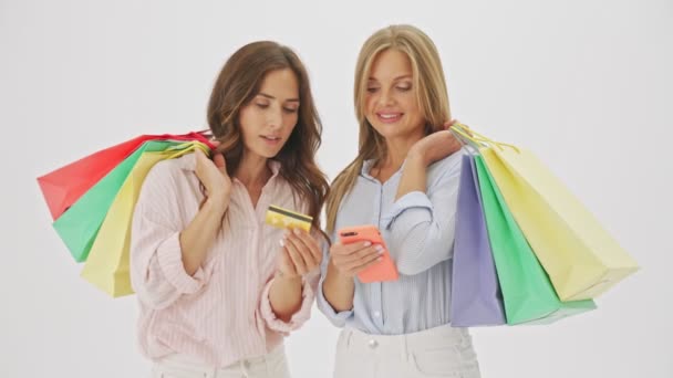 2人の素敵な陽気な女性はカラフルなショッピングバッグで立っており スタジオで白い背景に隔離されたスマートフォンやクレジットカードを使用しています — ストック動画