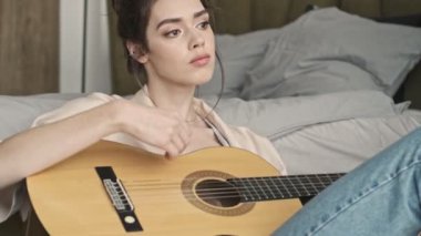 Evde konsantre olmuş genç bir kadın gitar çalarken yatağın yanında oturuyor.