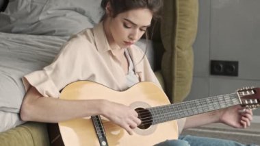 Evde konsantre olmuş genç bir kadın gitar çalarken yatağın yanında oturuyor.