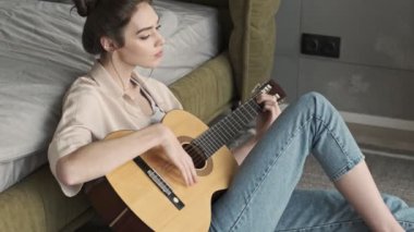 Evde konsantre olmuş genç bir kadın gitar çalarken yatağın yanında oturuyor.