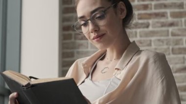 Genç, neşeli, iyimser kadın evde gözlükleriyle kitap okurken oturuyor.