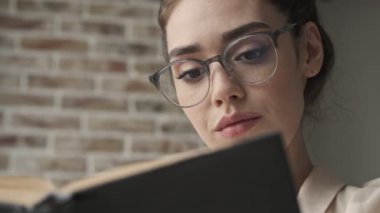 Evde gözlükleriyle kitap okurken konsantre olmuş genç bir kadın.