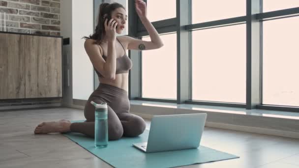 年轻而又集中的女运动员呆在室内的健身毯上 用手机和笔记本电脑聊天 — 图库视频影像