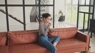Gülümseyen Asyalı güzel kadın akıllı telefon kullanıyor ve evdeki kanepede otururken çay içiyor.