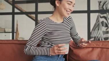 Akıllı telefon kullanan, çay içen ve evde kanepede otururken yüzünü çeviren düşünceli, güzel Asyalı kadın.