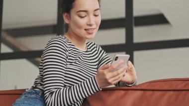 Evdeki kanepede otururken akıllı telefon kullanan güzel Asyalı kadın.