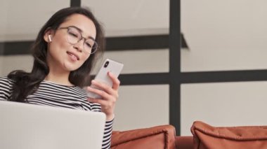 Evdeki kanepede dizüstü bilgisayarla otururken gözlüklü güzel Asyalı kadın akıllı telefon kullanıyor.