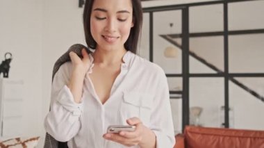 Gülümseyen Asyalı güzel bir kadın evde omzunda ceketle akıllı telefon kullanıyor.