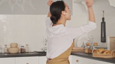 Neşeli, güzel Asyalı kadın önlük giyiyor kulaklıkla müzik dinliyor ve mutfakta dans ediyor.