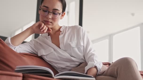 穏やかなエレガントなアジアの女性で眼鏡をかけ雑誌を読みながらソファに座って自宅 — ストック動画
