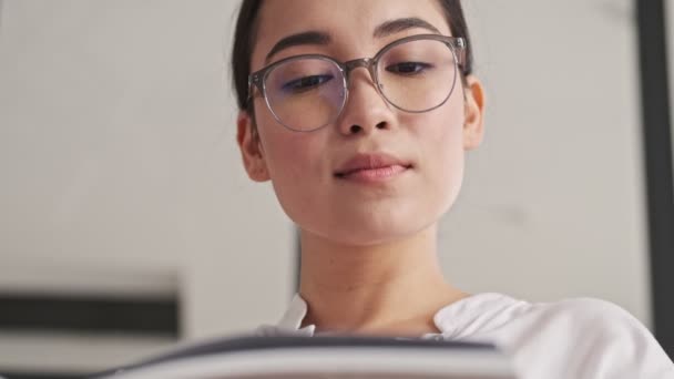 ケアフリーエレガントなアジア人女性で眼鏡をかけます雑誌を読みながらソファに家に座って — ストック動画