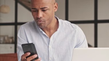 Gülümseyen Afro-Amerikan bir adam akıllı telefonuyla konuşuyor evde oturmuş dizüstü bilgisayarla çalışıyor.