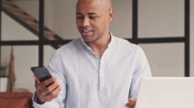 Yakışıklı bir Afrikalı Amerikalı akıllı telefonunu evde laptopuyla otururken kullanıyor.