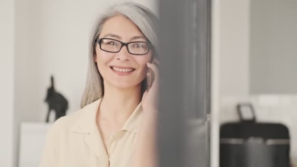 Uśmiechnięta Dojrzała Kobieta Okularach Rozmawia Przez Smartfona Stojącego Salonie Domu — Wideo stockowe