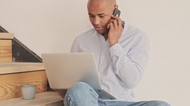 Çekici bir Afrikalı Amerikalı, evinde merdivenlerde otururken bilgisayarını kullanırken akıllı telefonuyla konuşuyor.